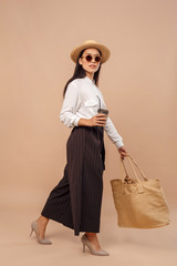 Freestyle. Young woman in shirt and sunglasses carrying bag walking isolated on bage with cup of coffee smiling confident full body shot