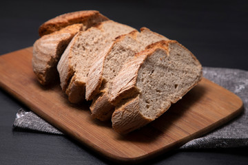 Freshly baked bread is cut into large pieces on a wooden board with a black background. Appetizing tasty bread close-up.