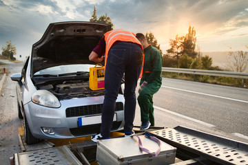 Two road assistant workers in towing service trying to start car engine with jump starter and...