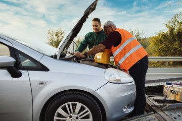 Two road assistant workers in towing service trying to start car engine with jump starter and...