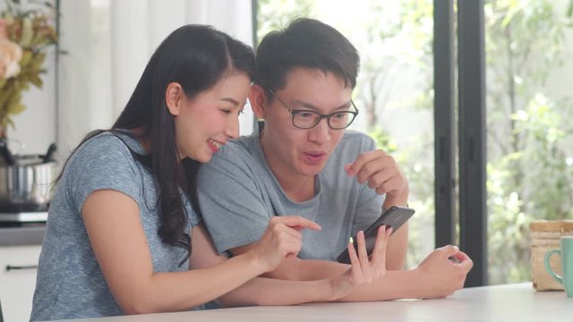 Young Asian couple enjoy shopping online on mobile phone at home. Lifestyle young husband and wife happy buy ecommerce after have breakfast in modern kitchen at house in the morning concept.