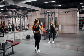 Obraz na płótnie Canvas Attractive cute fitness trainer wearing black sexy leggins and short t shirt, holding phone, taking selfie, standing with back towards camera, on background of mirror wall, in gym studio