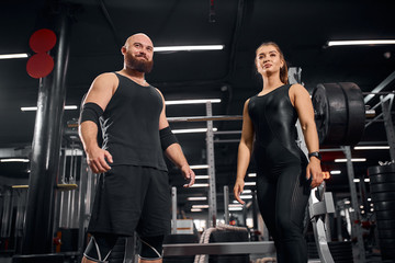 Cheerful happy powerlifters dressed in black sportswear, standing together after competition, looking away with glad expression, satisfied by good workout, low angle view, gym atmosphere