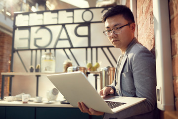 Side view portrait of modern Asian entrepreneur using laptop standing by brisk wall in office, copy space