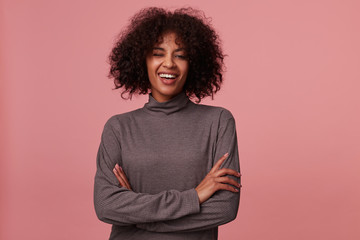 Fototapeta na wymiar Joyful attractive young curly brunette lady with dark skin keeping hands folded while looking cheerfully to camera and winking, standing against pink in casual wear