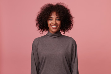 Fototapeta na wymiar Indoor shot of positive smiling young dark skinned woman with short curly brown hair looking happily to camera, standing over pink background with hands down