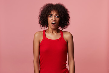Shocked young dark skinned lady with short brown curly hair wearing red shirt over pink background, keeping hands along her body and looking at camera with pout, rounding eyes with wide opened mouth