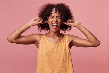 Fototapeta na wymiar Stressed dark skinned curly brunette female with short haircut covering her ears and screaming with wide mouth opened, frowning face with closed eyes while standing over pink background