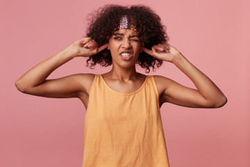 Fototapeta na wymiar Displeased young curly brunette female with dark skin wearing casual hairstyle over pink background, frowning her face with pout and inserting forefingers into her ears to avoid annoying sounds