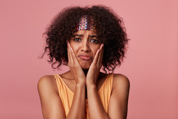 Unhappy brown-eyed young curly brunette female with dark skin looking sadly at camera and twisting her mouth, keeping palms on her cheeks while standing against pink background