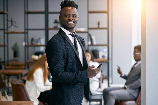 Personable businessman wearing formal black tuxedo stand posing in office environment and look at camera and smile . Using smartphone in working time. Business people concept