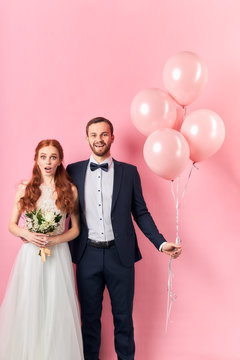 Happy smiling caucasian man in tuxedo hold pink air balloons and woman with opened mouth stand with bouquet isolated over pink background