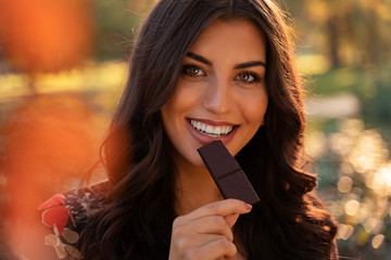 Gorgeous young woman taking the bite of dark chocolate outdoors at sunset. Smiling and looking at...