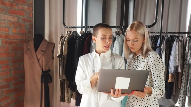 Two young caucasian women looks at laptop and enthusiastically speak about clothes. Background rack of clothes
