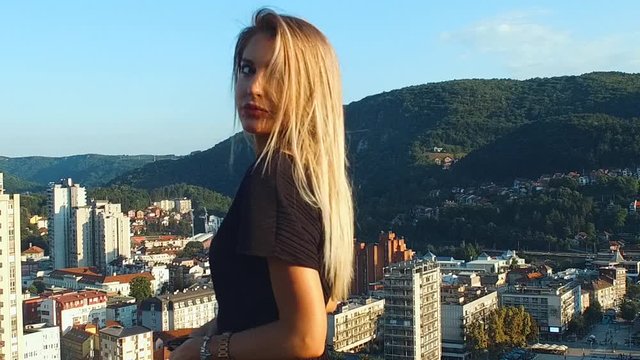 Attractive sexy fashionable young woman looking sensual over her shoulder, wind blowing in her blonde hair. Urban city background. Slow motion, evening sun.