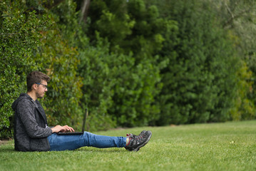 A blond man wearing glasses sitting on the grass of a park with his legs stretched. He is working with his laptop. Natural environment. Educational concept