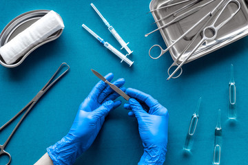 Preparing for plastic surgery. Doctor's hands takes scalpel on blue background with surgical tools top view