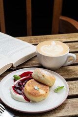 Cheese pancakes with sour cream and cherry sauce and cappuccino on a wooden table with book