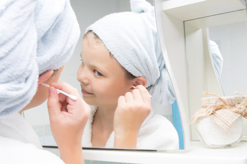 Obraz na płótnie Canvas a girl in the bathroom, in a white bathrobe and a towel on her head, cleans her ears with a cotton swab, looking in the mirror