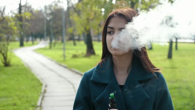 Cute caucasian brunette businesswoman in coat relaxing by smoking electronic cigarette in the green park in the autumn