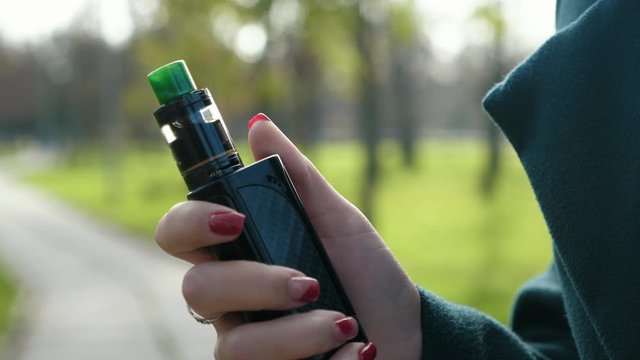Cool black box mod device for vaping with green drip tip in hand of young girl on the street in a park, Slow motion