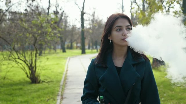 Beautiful young brunette girl walking in the park with green grass and vaping, Slow motion