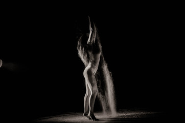 Fototapeta na wymiar The girl with the flour on the body stretches the arms up with thrown flour on black background black and white image