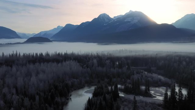 Aerial: sunrise view over forest, rocky mountains and the Blue River. Fog is covering the forest and it feels tranquil. Blue River, British Columbia, Canada