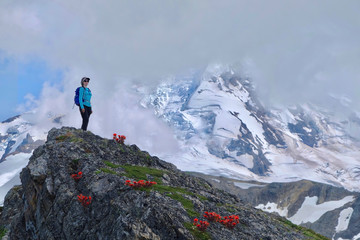 Middle age  woman in sport clothes with a backpack standing on hill  with red flowers by a mountain covered with glaciers and snow in clouds. Indian Paintbrush on rocks near Mt Baker. WA. USA 