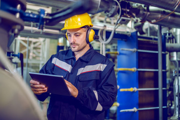 Dedicated factory worker standing next to boiler and holding tablet. Worker is dressed in protective uniform, having hardhat and antiphons.