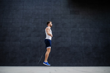 Fototapeta na wymiar Side view of handsome muscular caucasian man in shorts and t-shirt skipping rope in front of gray wall outdoors.