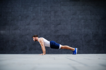Fototapeta na wymiar Side view of handsome Caucasian muscular bearded man in shorts and t-shirt doing push-ups. In background is gray wall.