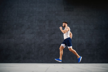Fototapeta na wymiar Side view of handsome caucasian muscular fit man running fast outdoors. In background is gray wall.