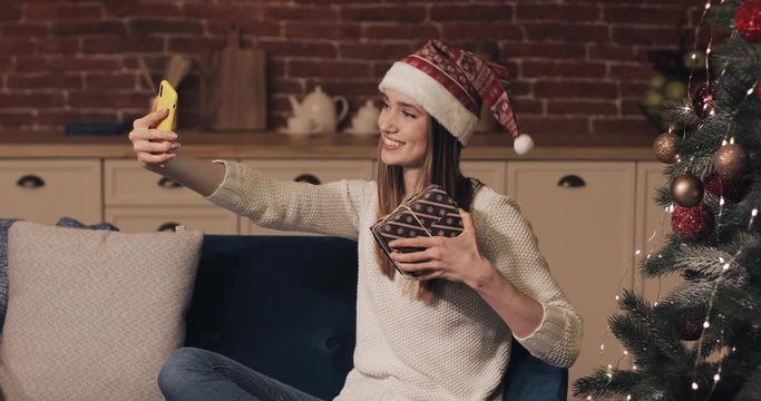 Happy Caucasian Young Pretty Girl Wearing Christamas Hat Sitting on the Sofa Holding and Shaking Present Box near Christmas Tree Making a Video Call or Video Message.