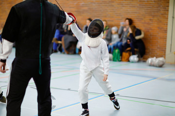 Little kid boy fencing on a fence competition. Child in white fencer uniform with mask and sabre....