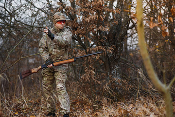 A male hunter with a gun while sitting takes aim at a forest. The concept of a successful hunt, an experienced hunter. Hunting the autumn season. The hunter has a rifle and a hunting uniform.