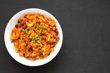 Homemade Chili Mac and Cheese with Parsley in a white bowl over black background, top view. Flat lay, overhead, from above. Copy space.