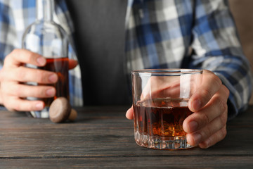 Man in shirt holds bottle and glass of whiskey on wooden table, close up