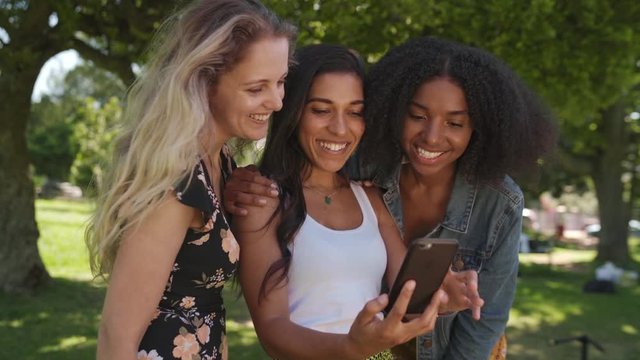 Happy young group of multi ethnic female friends browsing using smartphone technology enjoying sharing moments with each other in the park on a sunny day