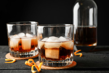 Glasses of whiskey with orange peels and ice on wooden background with bottle, close up