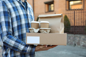 Man holds blank box, coffee cups and paper package outdoor. Delivery