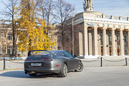 A rare Japanese sports car in the back of a gray Toyota Supra coupe with a spoiler on the road in city near trees with yellow leaves and an old building with columns.