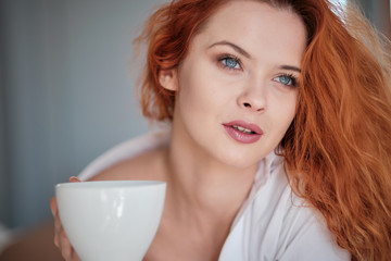 Obraz na płótnie Canvas Attractive redhaired woman with a cup of coffee on the bed