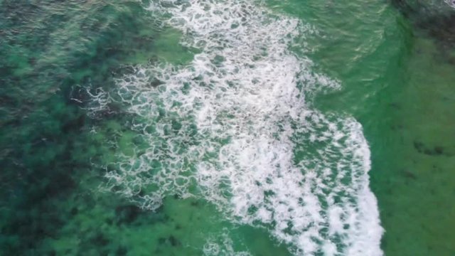 Aerial Drone Above Crystal Clear Blue Ocean Water On A Sunny Day With Rocky Sandy Bottom With Crashing White Wash Waves