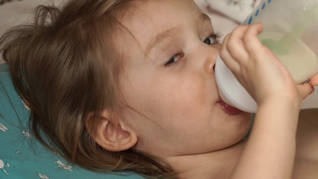Portrait of little baby girl lies on bed after sleep, holds bottle of milk, hands and sucks infant formula milk with great appetite. Baby food. Сlose up