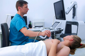 Man doctor making ultrasound diagnostic of mammary glands of young woman on bust. He runs ultrasound sensor over patient's mammary glands and looks at image on screen. Diagnosis of internal organs.