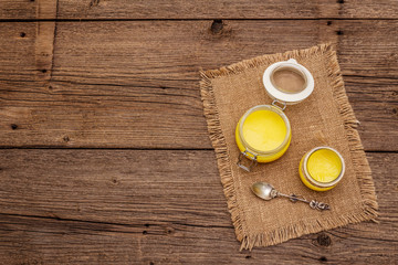 Pure or desi ghee (ghi), clarified melted butter. Healthy fats bulletproof diet concept or paleo style plan. Glass jar, silver spoon on vintage sackcloth. Wooden boards background