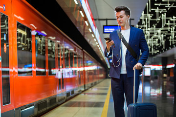 Young stylish handsome man in suit with suitcase standing on metro station holding smart phone in hand, scrolling and texting, smiling and laughing. Futuristic bright subway station. Finland