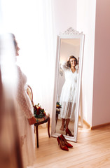 Bride in elegant white dress smiles and poses in front of the mirror.  Beautiful bride is having fun and preparing for her wedding. Beautiful woman with  make up and hair style. Morning of the bride.