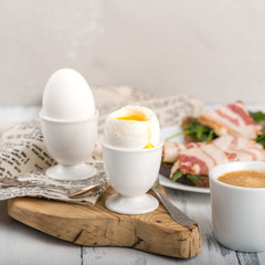 Two boiled eggs, in a shell, a drop of yolk, in a white egg stand, on a wooden Board, a textile napkin, toast with bacon on a plate, a Cup of coffee, light gray background